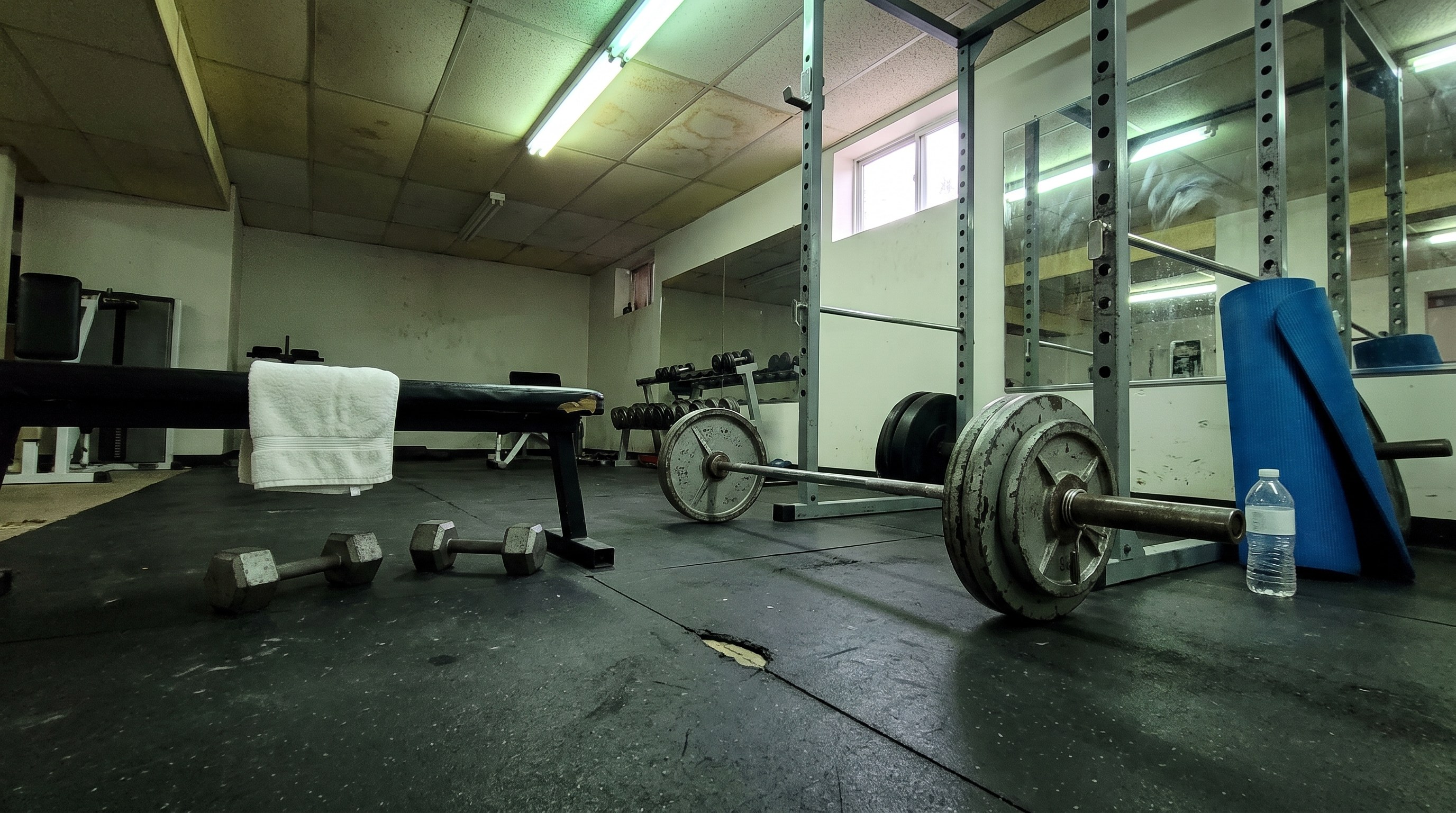 A loaded barbell and a chipped power rack on the scuffed rubber floor of an ordinary strip mall gym